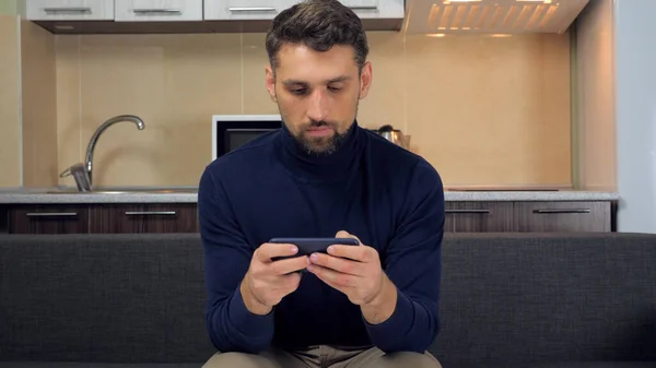 Bearded male gamer sits on couch at home plays video game on smartphone — Stock Photo, Image