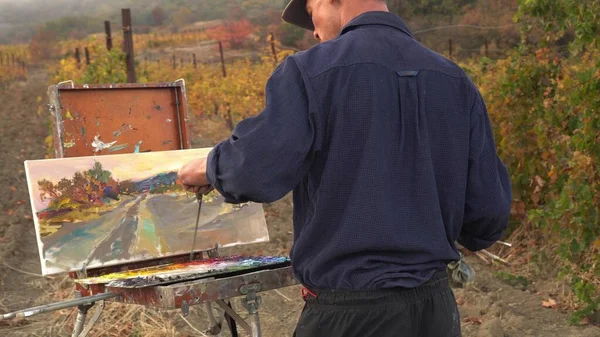 Professional artist man paints a picture on canvas using oil paints. Landscape Oil Painting. Autumn vineyard