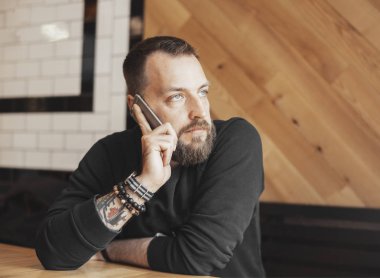 Kafede telefonla konuşan sakallı çekici dövmeli adam.
