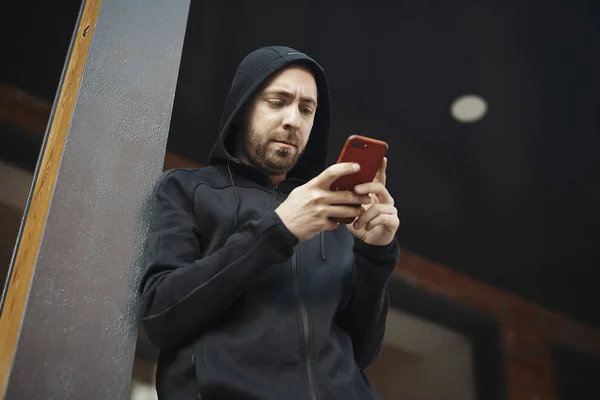 Hombre barbudo de pie junto a poste de hormigón y mirando la pantalla . — Foto de Stock