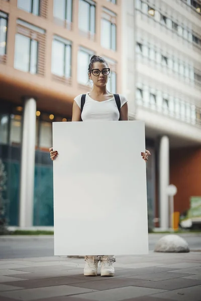 Mladá atraktivní brýlatý žena držící prázdné plátno na ulici. — Stock fotografie