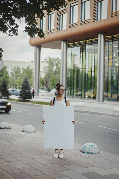Молодая привлекательная женщина держит чистый холст на улице и смотрит в сторону
.