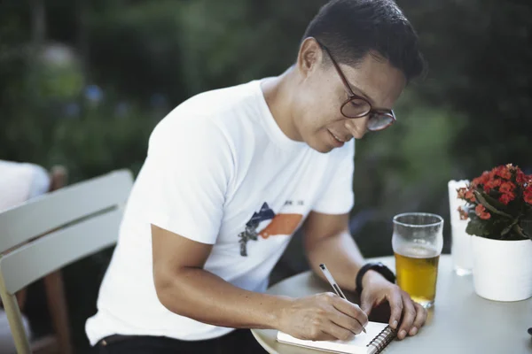 Jeune homme à lunettes buvant de la bière froide et écrivant quelques notes . — Photo