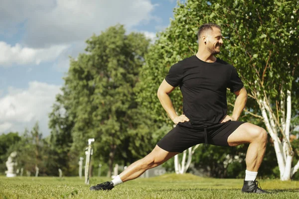 Jeune homme séduisant en tenue de sport noire faisant fente en plein air dans le parc . — Photo