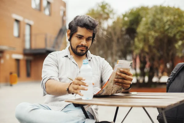 Hombre atractivo sentado en la cafetería de la calle, mirando el mapa y sosteniendo la botella . — Foto de Stock