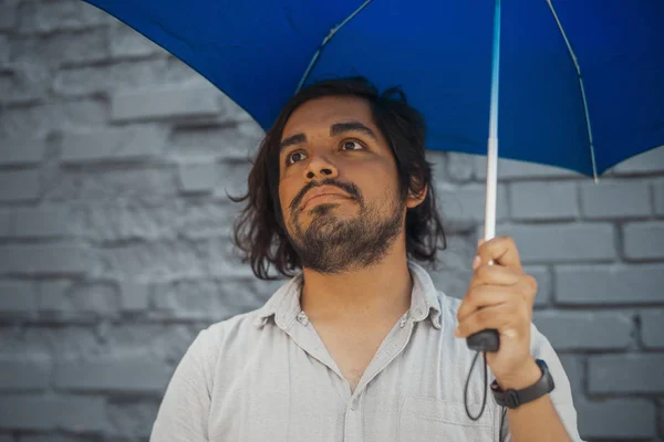 Attractive brunette latin man holding blue umbrella next to grey brick wall.