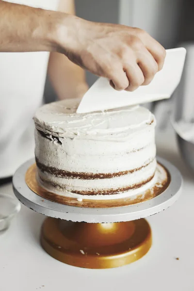 Close up of hands smearing butter cream on chocolate cake by white spatula. — Stock Photo, Image
