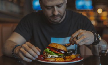 Genç çekici adam yeme hamburger ve patates kızartması kafe yakın çekim.