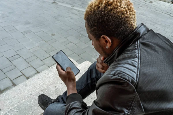 Hombre de piel oscura mirando la pantalla del teléfono móvil y sentado en escalones concretos . — Foto de Stock