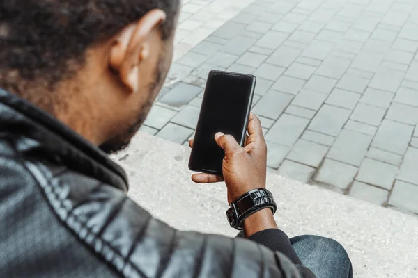 Hombre de piel oscura mirando la pantalla del teléfono móvil y sentado en escalones concretos . — Foto de Stock