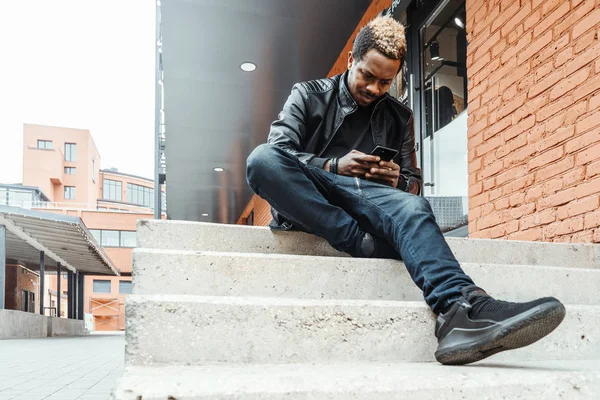 Hombre sonriente de piel oscura mirando la pantalla del teléfono móvil y sentado en los escalones . — Foto de Stock