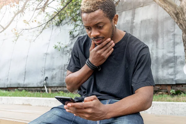 Mietteliäs tummaihoinen mies mustassa T-paidassa katselemassa matkapuhelimen näyttöä . — kuvapankkivalokuva