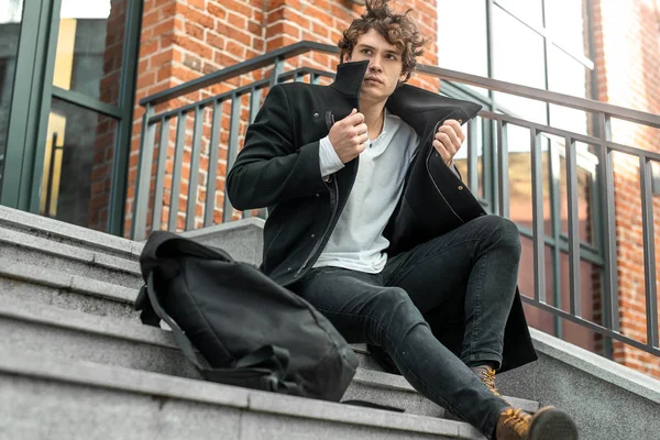 Homme pensif assis sur des marches en béton et enveloppant dans un manteau noir — Photo