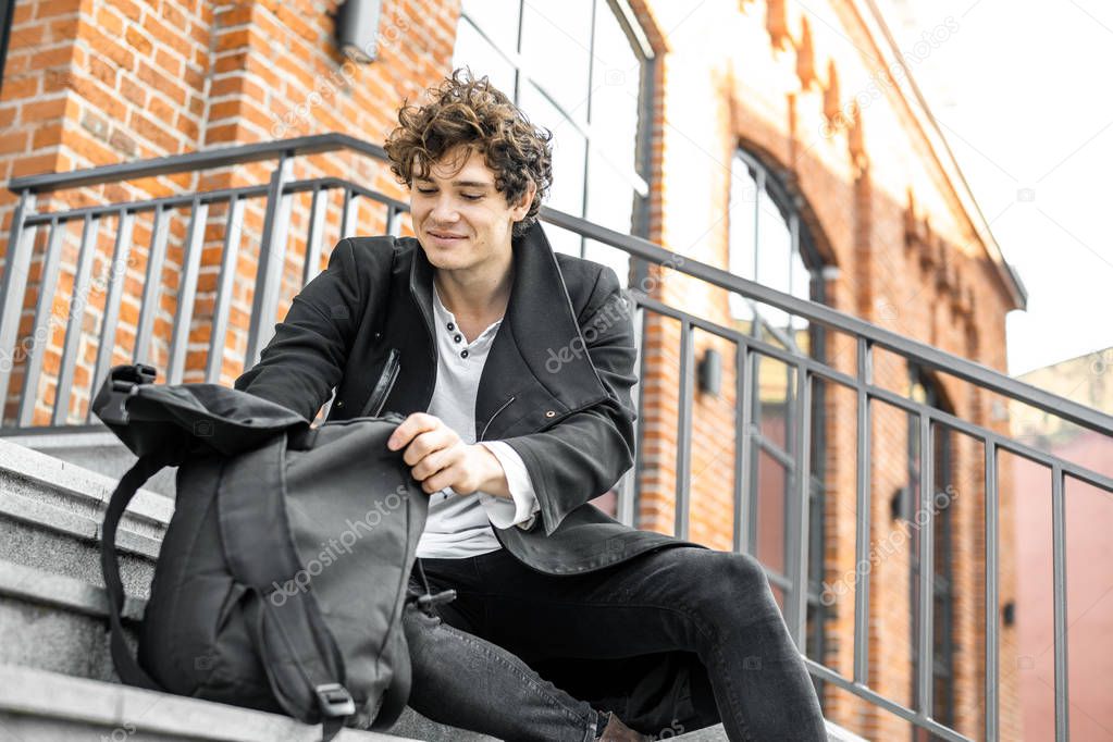 Young attractive pensive man sitting on steps and rummaging in his backpack.