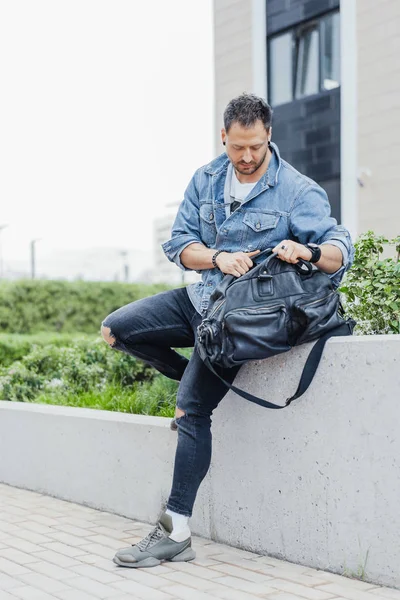 Joven buscando en bolsa de deporte negro y sentado en el fondo del paisaje urbano . — Foto de Stock