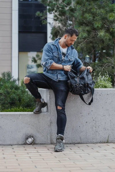 Joven buscando en bolsa de deporte negro y sentado en el fondo del paisaje urbano . — Foto de Stock