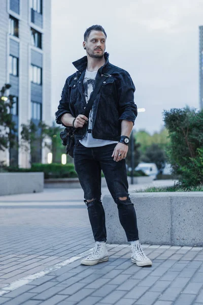Joven hombre atractivo en chaqueta vaqueros negro de pie en el fondo del paisaje urbano . — Foto de Stock