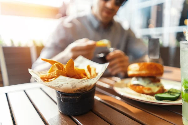 Close up of young attractive man eating french fries and burger at street cafe.