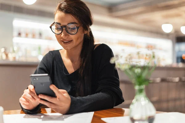 Ung vacker kvinna som håller mobiltelefon i händer i väntan på hennes måltid. — Stockfoto