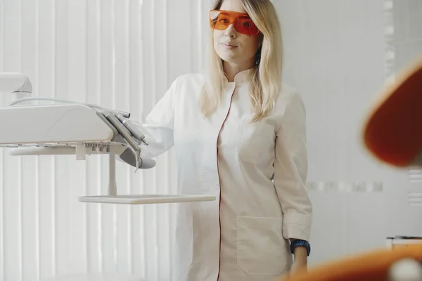 Блондинка-дантист в белой униформе, стоящая рядом с стоматологическим оборудованием . — стоковое фото