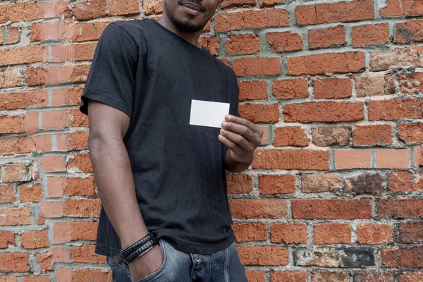 Dunkelhäutiger Mann im T-Shirt mit leerer Visitenkarte auf gemauertem Hintergrund. — Stockfoto