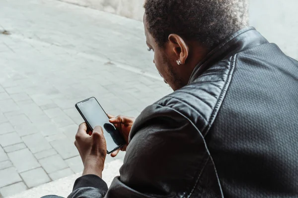 Dark-skinned man looking at mobile phone screen and sitting on concrete steps.