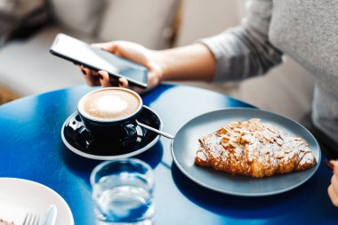 Kadın el fincan kahve, kruvasan, su ve cep telefonu ile tablo tutarak yakın çekim.