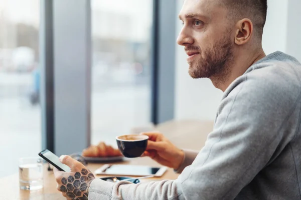 Primer plano del hombre con tatuaje sosteniendo taza de café y teléfono móvil en la cafetería . — Foto de Stock