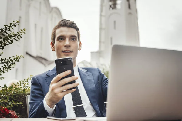 Młody biznesmen, siedzący obok laptopa i trzyma w ręku telefon komórkowy. — Zdjęcie stockowe