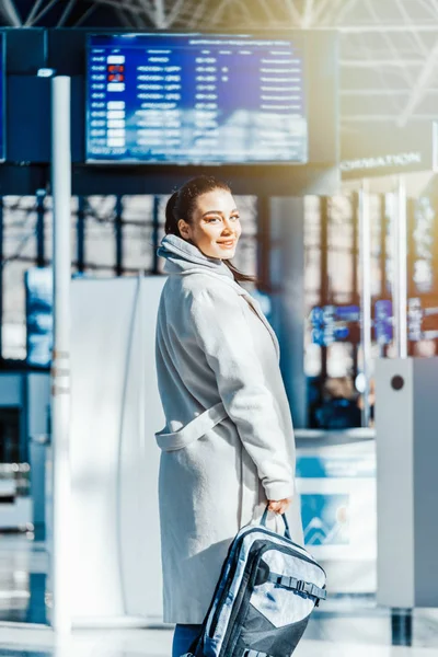 Mujer joven de pie frente a la pizarra de destino y sonriendo en el aeropuerto . —  Fotos de Stock