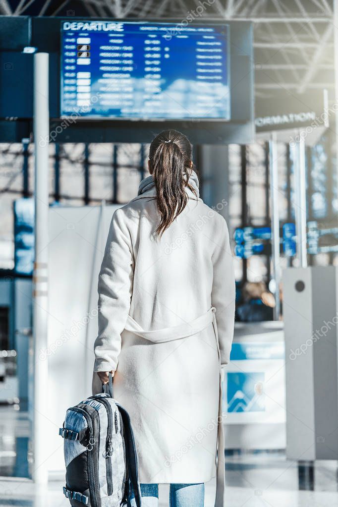 Young brunette woman standing in front of departure and arrival flights digital board in modern airport and checking her flight data. Destination screen board. happy vacation and travel concept.