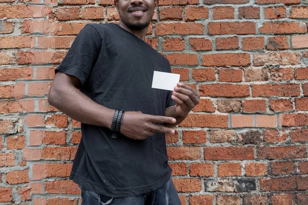 穿 t恤衫的黑皮肤男子在砖砌的背景上显示空白名片. — 图库照片
