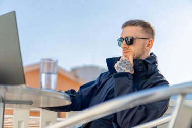 Yakışıklı, düşünceli, stil sahibi adam balkonda oturuyor ve dizüstü bilgisayarla çalışıyor..