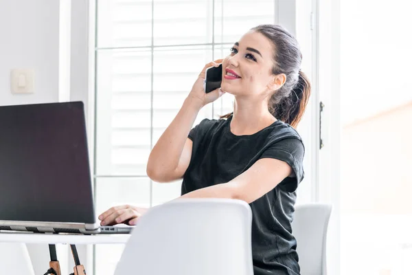 Fiatal nő dolgozik laptop és beszél mobiltelefonnal fényes lakások. — Stock Fotó