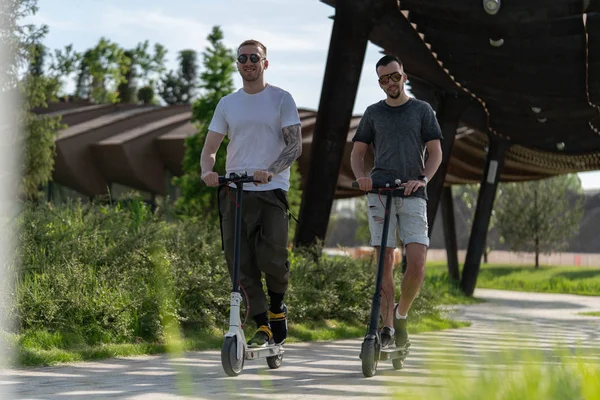 Zwei gutaussehende Männer fahren Elektro-Tretroller in schöner Parklandschaft — Stockfoto