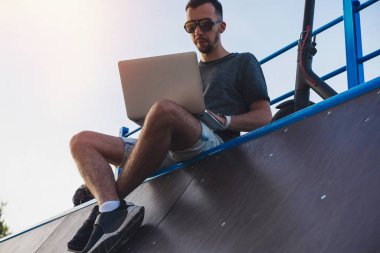 Modern parkta patenci rampasında oturan dizüstü bilgisayar ve elektrikli tekme scooter ile Adam.