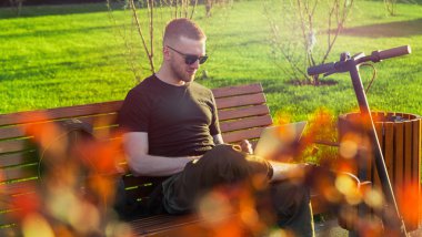 Çağdaş parkta oturan dizüstü bilgisayar ve modern elektrikli tekme scooter ile Man.