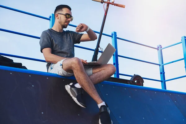 Modern parkta patenci rampasında oturan dizüstü bilgisayar ve elektrikli tekme scooter ile Adam.