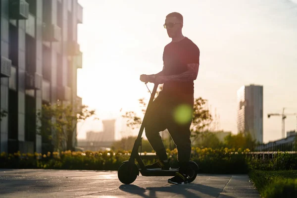 Homem montando scooter elétrico preto pontapé no fundo cityscape ao pôr do sol — Fotografia de Stock