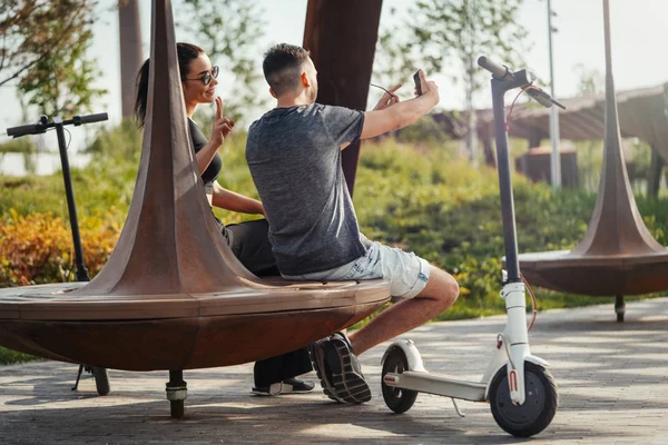 Pár fiatal férfi és nő ül a parkban, és így selfie. — Stock Fotó