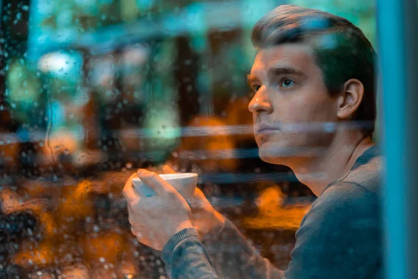 Yakışıklı adam yağmurlu bir günde çay veya kahve içiyor ve pencereden bakıyor. — Stok fotoğraf