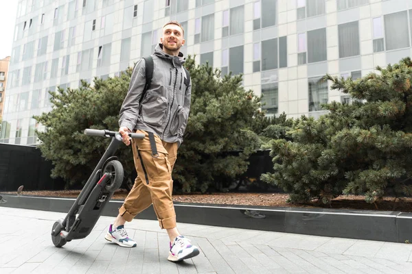 Unga attraktiva leende man som innehar elektrisk Spark skoter medan du går på en promenad på gatan. — Stockfoto