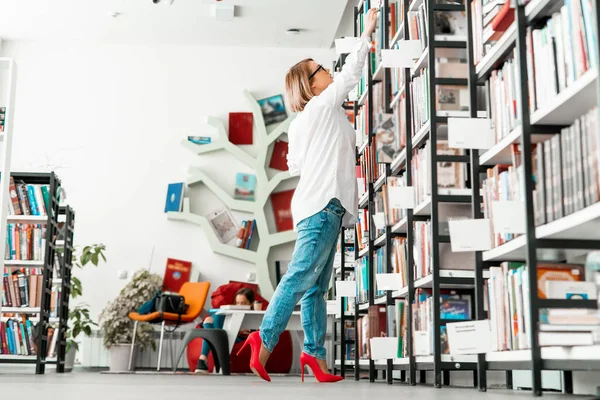 Młoda atrakcyjna kobieta, która ciągnie rękę do książki w sklepie z książką lub w bibliotece — Zdjęcie stockowe