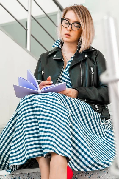 Молодая женщина делает заметки в фиолетовом блокноте в библиотеке и сидит на лестнице — стоковое фото