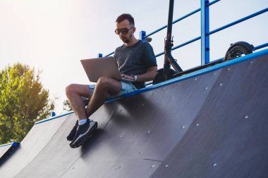 Modern parkta patenci rampasında oturan dizüstü bilgisayar ve elektrikli tekme scooter ile Adam.