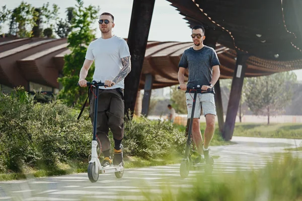 Zwei gutaussehende Männer fahren Elektro-Tretroller in schöner Parklandschaft — Stockfoto