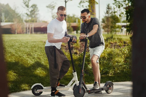 Двоє красивих чоловіків розмовляють поруч з електричними скутерами на парковому ландшафті — стокове фото