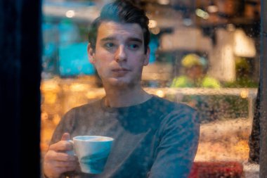 Genç dalgın yakışıklı adam rahat modern kafede yağmurlu bir günde çay veya kahve içme ve pencereden bakarak.