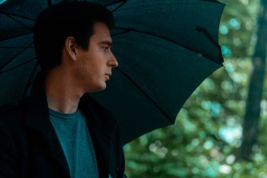 Yağmurlu günde şemsiyeli genç dalgın yakışıklı adam parkta yürüyüşe çıkıyor.