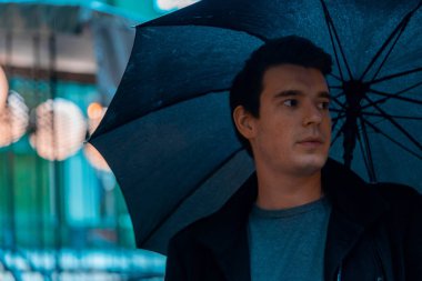 Yağmurlu günde şemsiyeli genç dalgın yakışıklı adam parkta yürüyüşe çıkıyor.